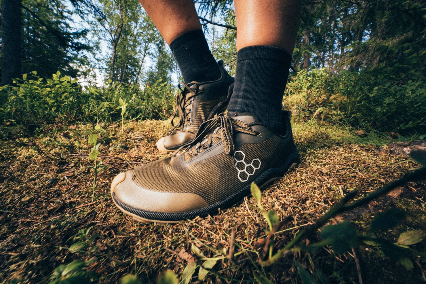 A close-up outdoor shot of a person's legs and feet wearing brown hiking boots with black socks, standing on a forest floor covered in pine needles and green plants.
