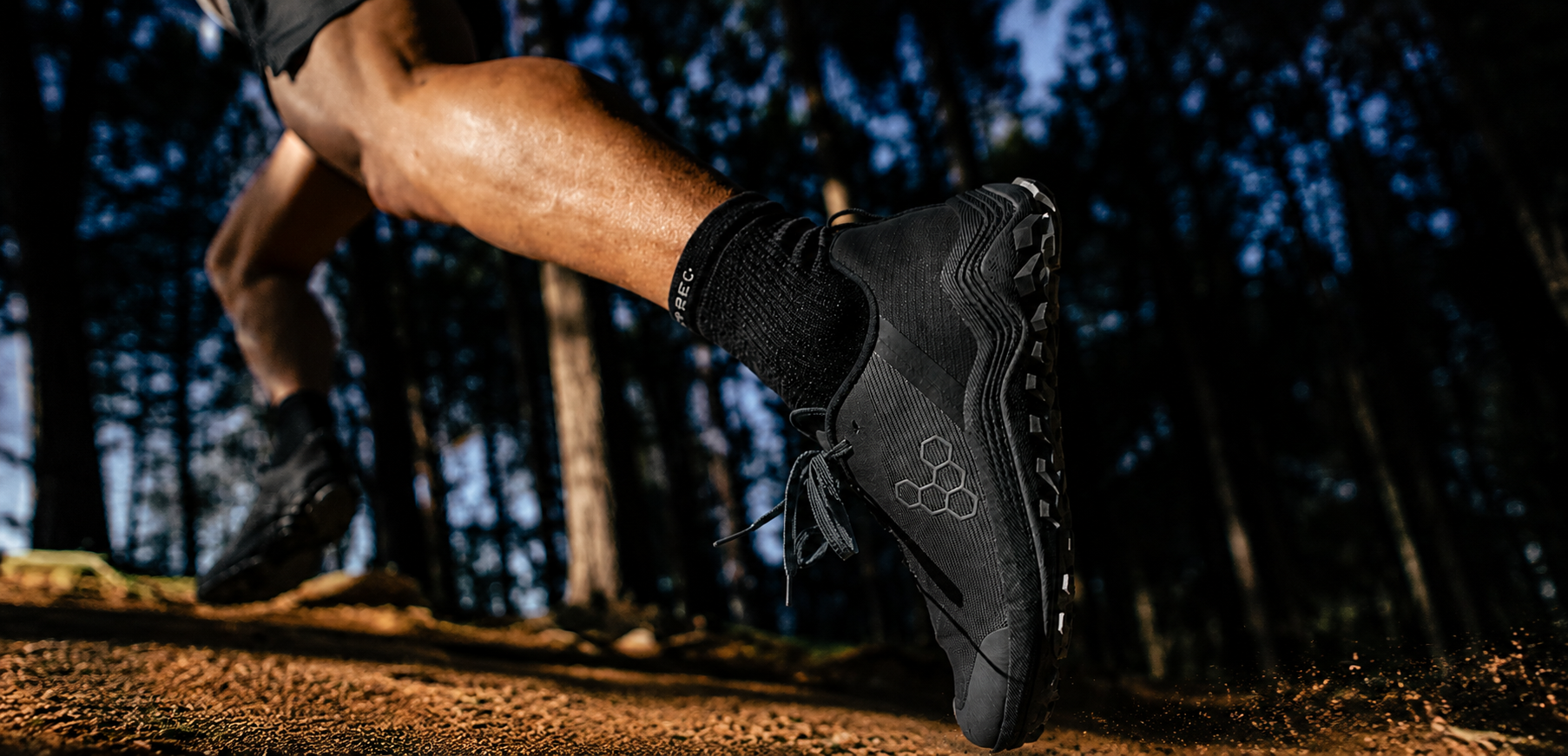 A close-up action shot of a runner's leg and foot wearing a black trail running shoe on a dirt trail in a forest.
