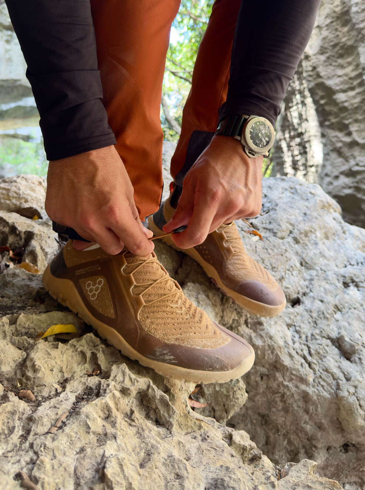 A person crouches on a rocky, uneven surface while adjusting the laces of brown and tan Vivobarefoot Primus Trail Knit FG Grounding shoes.