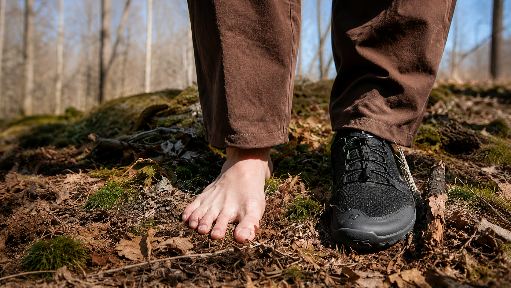 A close-up outdoor shot of a person's bare foot and right foot wearing a black hiking shoe, standing on a forest floor covered in leaves and moss.