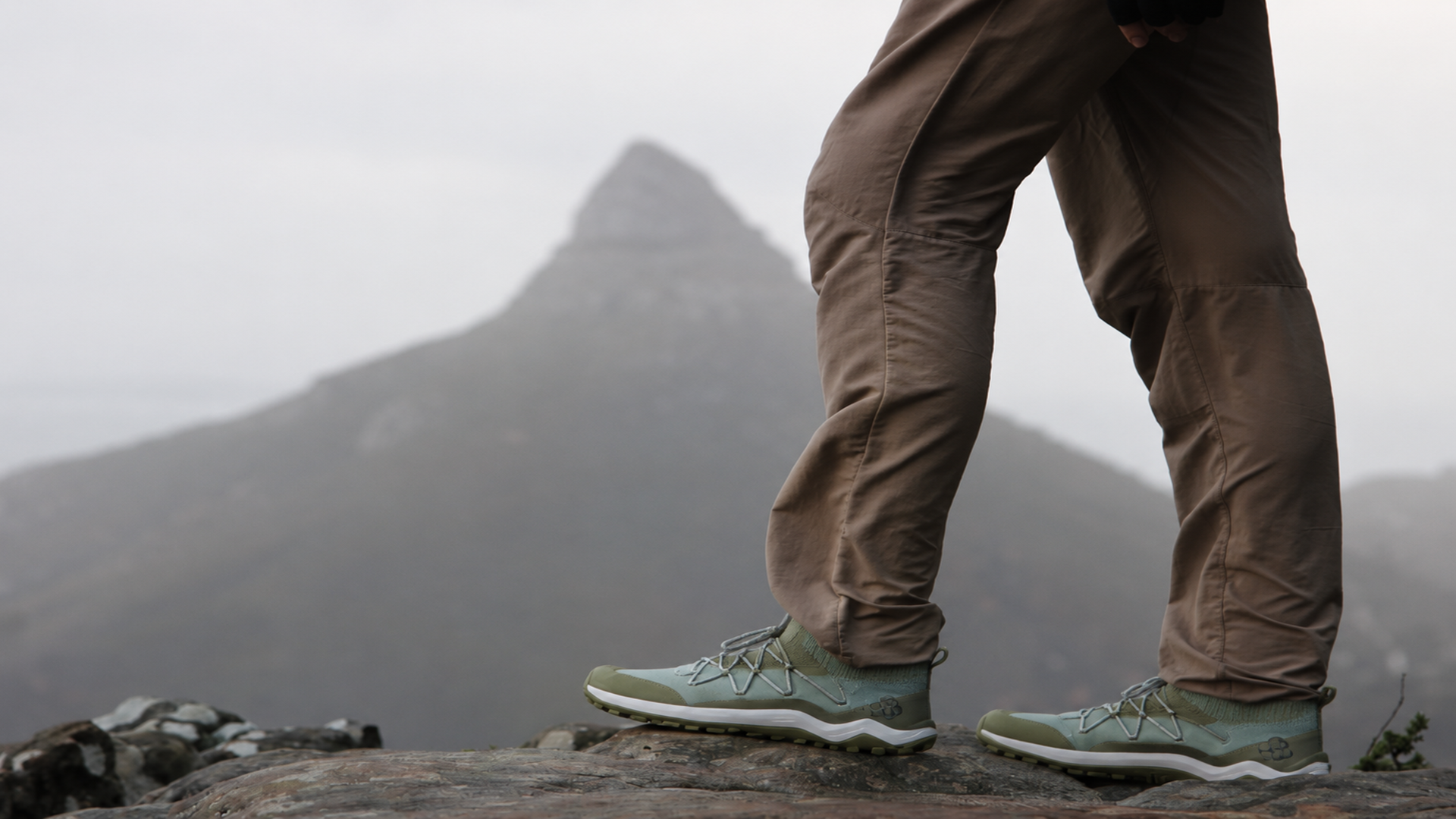 Person wearing Vivobarefoot hiking shoes standing on a rocky mountain trail with misty peaks in the background.