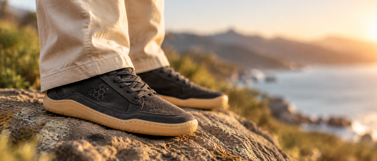 Vivobarefoot black suede barefoot sneakers with natural gum soles, worn on a rocky coastal trail in warm summer sunlight with a softly blurred ocean background.