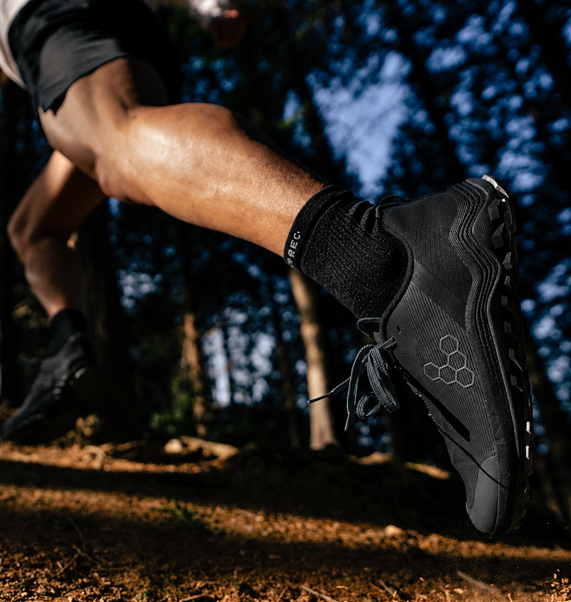 A close-up action shot of a person running outdoors in a forest, wearing black athletic shoes and black socks. The shoe has a visible hexagonal logo pattern on the side.
